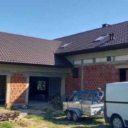 Bud-Henryk - Dom jednorodzinny w trakcie budowy z nowym brązowym dachem i elewacją z czerwonej cegły, widoczny pracownik z przyczepą i biały samochód dostawczy.