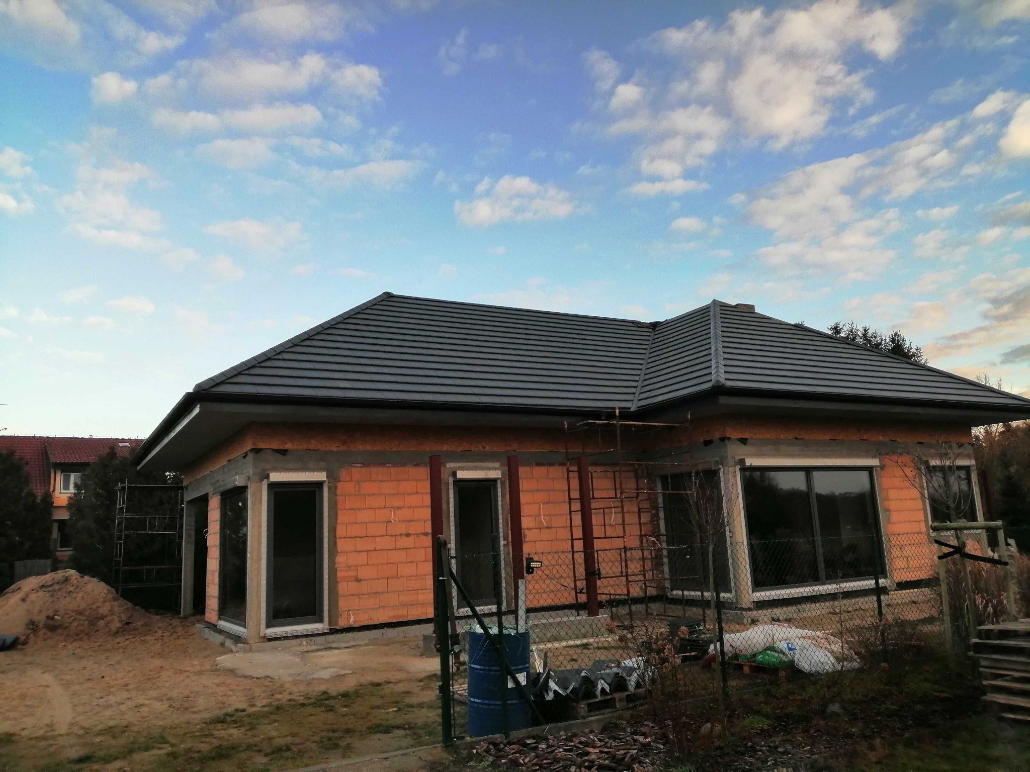 Dom jednorodzinny w trakcie budowy z widoczną nową, grafitową dachówką, surowymi ścianami z czerwonej cegły i zamontowanymi oknami, na tle błękitnego nieba z obłokami.