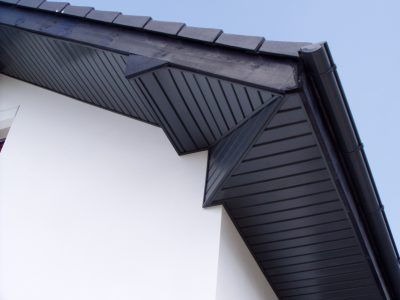 Detale wykończenia dachu z ciemną podbitką i rynną na tle białej ściany budynku, widoczne dachówki i fragment nieba.