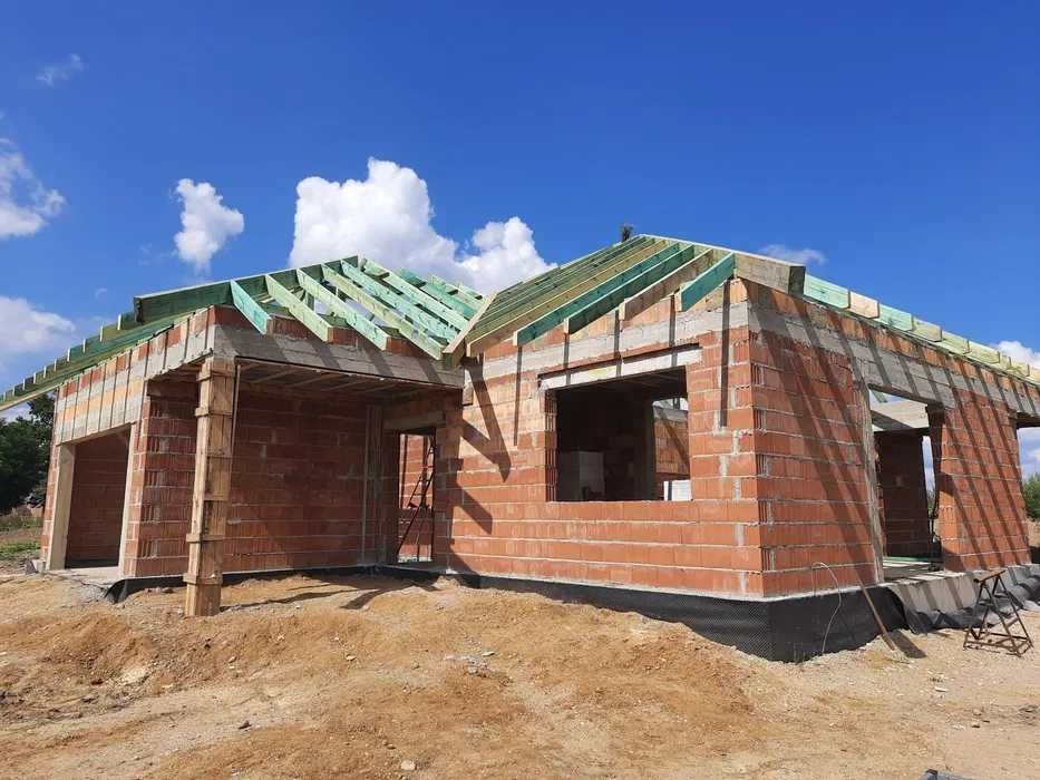 Surowy stan domu jednorodzinnego z czerwonej cegły, widoczne elementy konstrukcji dachu z zielonej impregnacji, czarne zabezpieczenie fundamentów, ziemisty teren budowy i błękitne niebo z chmurami...