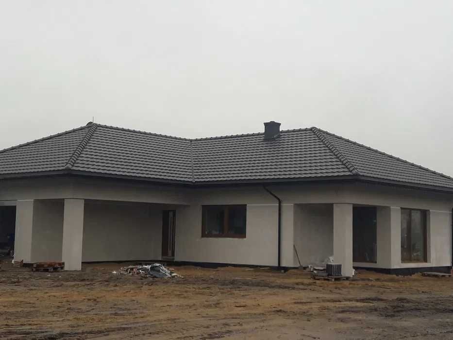 Nowoczesny dom jednorodzinny z szarą dachówką karpiówką i jasną elewacją, widoczne rynny i komin, teren wokół domu w trakcie zagospodarowywania.