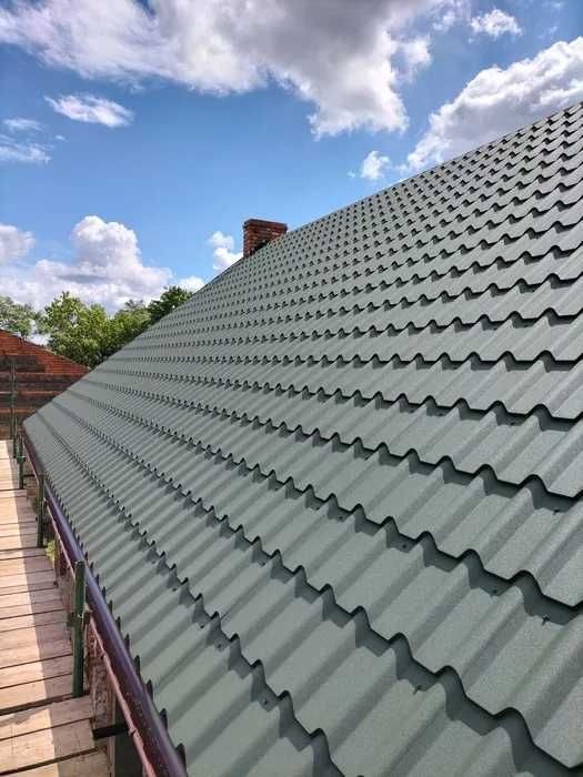 Szaro-zielony dach z blachodachówki modułowej, widoczny z poziomu rusztowania, z kominem w tle i błękitnym niebem z obłokami.