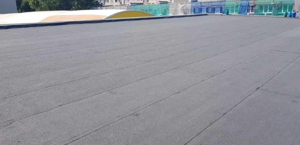 Szary, płaski dach pokryty papą termozgrzewalną z widocznymi łączeniami, na tle częściowo widocznych budynków.