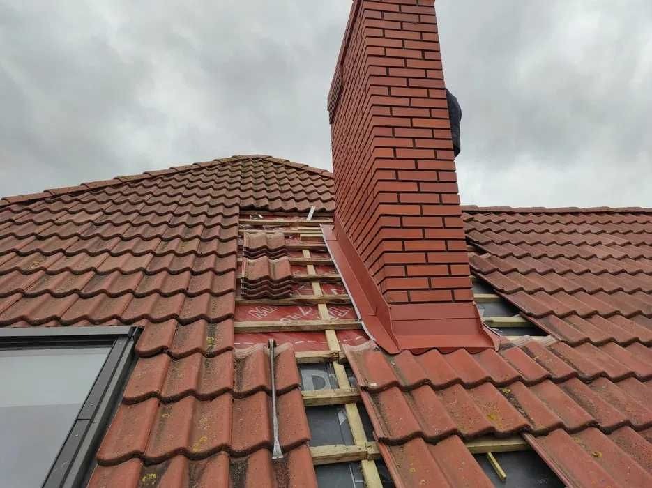 Częściowo zdjęte dachówki na dachu z widoczną konstrukcją drewnianą i kominem z cegły, narzędzie dekarskie leżące na dachu, pochmurne niebo w tle.