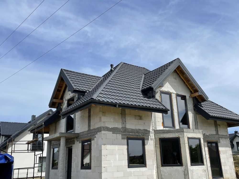 Dom w stanie surowym zamkniętym we Wrocławiu z grafitowym dachem z blachodachówki i czarnymi oknami, widok z dołu na tle błękitnego nieba.