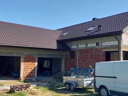 Dom jednorodzinny w trakcie budowy z nowym brązowym dachem i elewacją z czerwonej cegły, widoczny pracownik z przyczepą i biały samochód dostawczy.