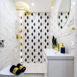 Nowoczesna łazienka z białymi marmurkowymi płytkami, kabiną prysznicową z czarno-białymi rombami, złotą armaturą, okrągłym lustrem i białą szafką pod umywalką. Na toalecie leżą zwinięte, granatowe...