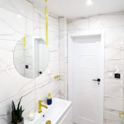 Nowoczesna łazienka z białymi marmurkowymi płytkami, złotą armaturą, okrągłym lustrem i białą szafką umywalkową z rośliną w szarej doniczce.