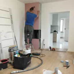 Mężczyzna na drabince montuje płytę kartonowo-gipsową na ścianie z przygotowaną konstrukcją metalową, widoczny od tyłu, w tle otwarte przejście do kuchni, na podłodze narzędzia i akcesoria...