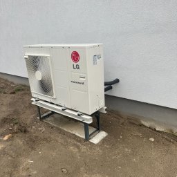Proxet Jacek Wieczorkowski - Biała pompa ciepła LG Therma V zamontowana na metalowej konstrukcji przy ścianie budynku. Widoczne rury i fragment terenu.