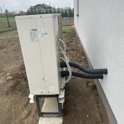 Proxet Jacek Wieczorkowski - Boczny widok zainstalowanej pompy ciepła LG na zewnątrz budynku z rurami wchodzącymi w ścianę. Widoczne elementy montażowe i otoczenie.