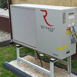 Proxet Jacek Wieczorkowski - Pompa ciepła Rotenso na zewnątrz budynku, zamontowana na metalowej konstrukcji, otoczona kamieniami i obrzeżem. Widoczna ściana budynku i zieleń.