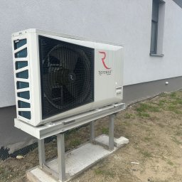 Proxet Jacek Wieczorkowski - Biała pompa ciepła Rotenso na metalowej konstrukcji przy szarej ścianie budynku. Widoczne logo producenta i fragment okna. Tło: trawa i ogrodzenie.