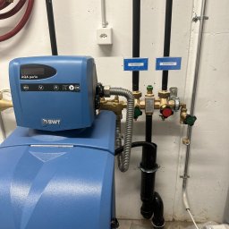 Proxet Jacek Wieczorkowski - Niebieski zmiękczacz wody BWT AQA Perla z podłączonymi rurami, zaworami i oznaczeniami 'Kaltwasser' i 'Warmwasser' w kotłowni lub pomieszczeniu technicznym.