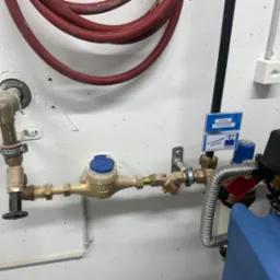 Zamontowany wodomierz z rurami, zaworami i przyłączami na białej, teksturowanej ścianie. Widoczny przewód i etykiety 'Garten' oraz logo firmy instalacyjnej.