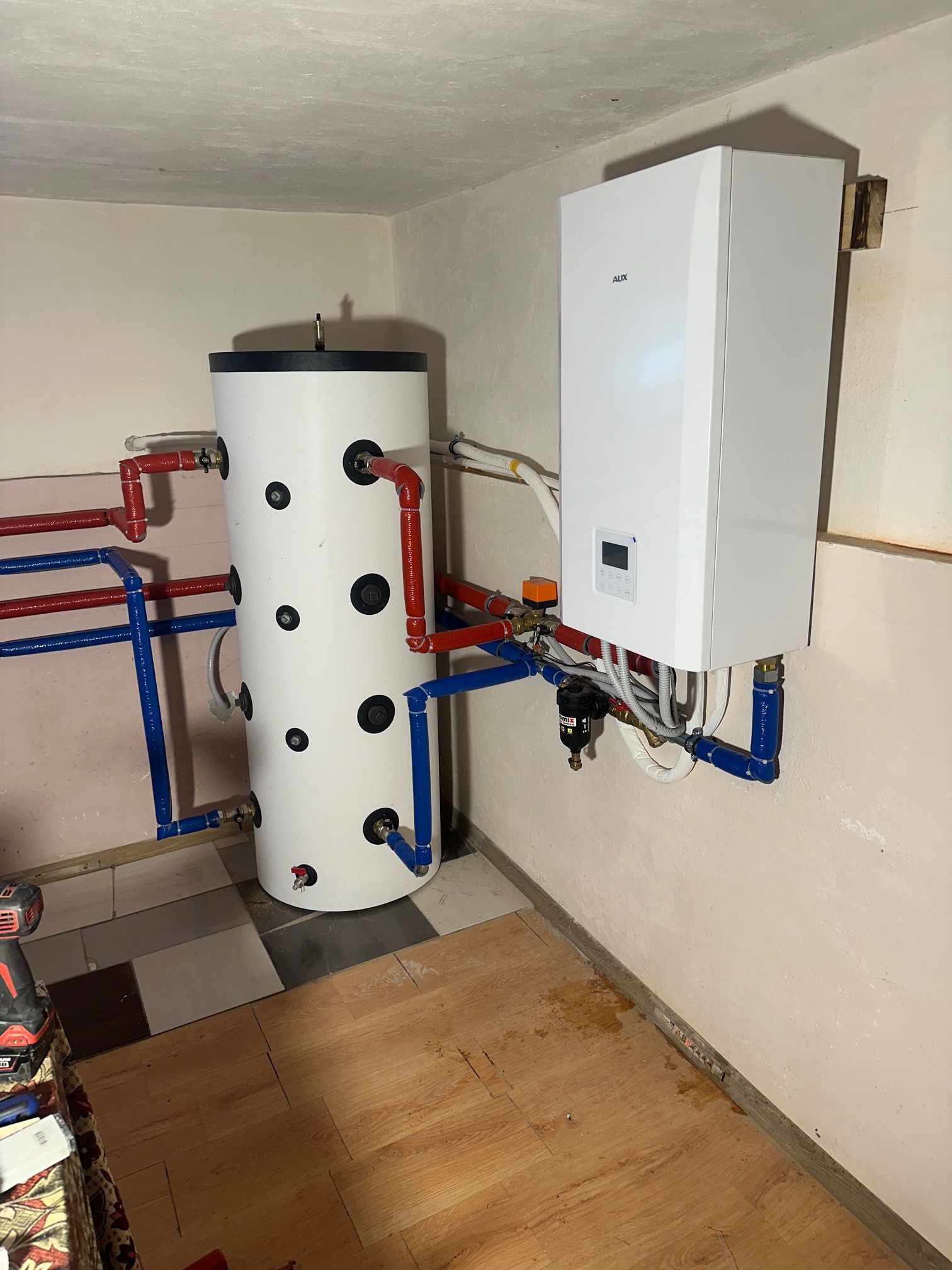 Biały zasobnik ciepłej wody z czarnymi okręgami i piec gazowy AUX na ścianie, połączone rurami w kolorach czerwonym i niebieskim, w kotłowni z brudną podłogą.