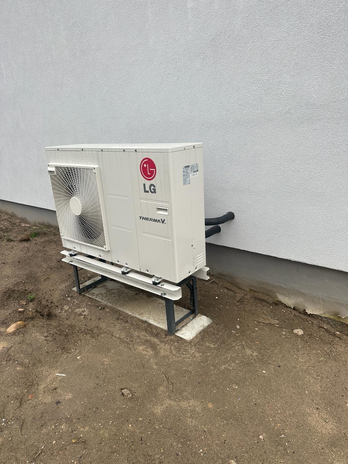 Biała pompa ciepła LG Therma V zamontowana na metalowej konstrukcji przy ścianie budynku. Widoczne rury i fragment terenu.