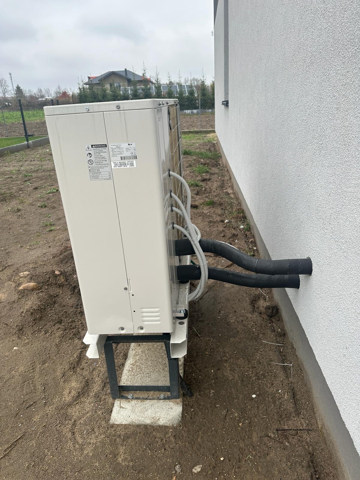 Boczny widok zainstalowanej pompy ciepła LG na zewnątrz budynku z rurami wchodzącymi w ścianę. Widoczne elementy montażowe i otoczenie.