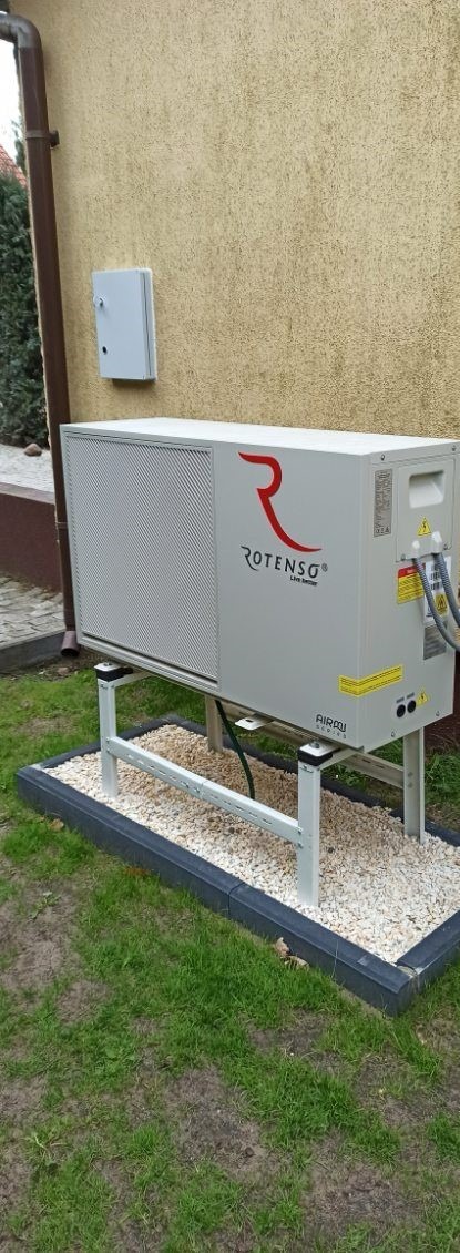 Pompa ciepła Rotenso na zewnątrz budynku, zamontowana na metalowej konstrukcji, otoczona kamieniami i obrzeżem. Widoczna ściana budynku i zieleń.