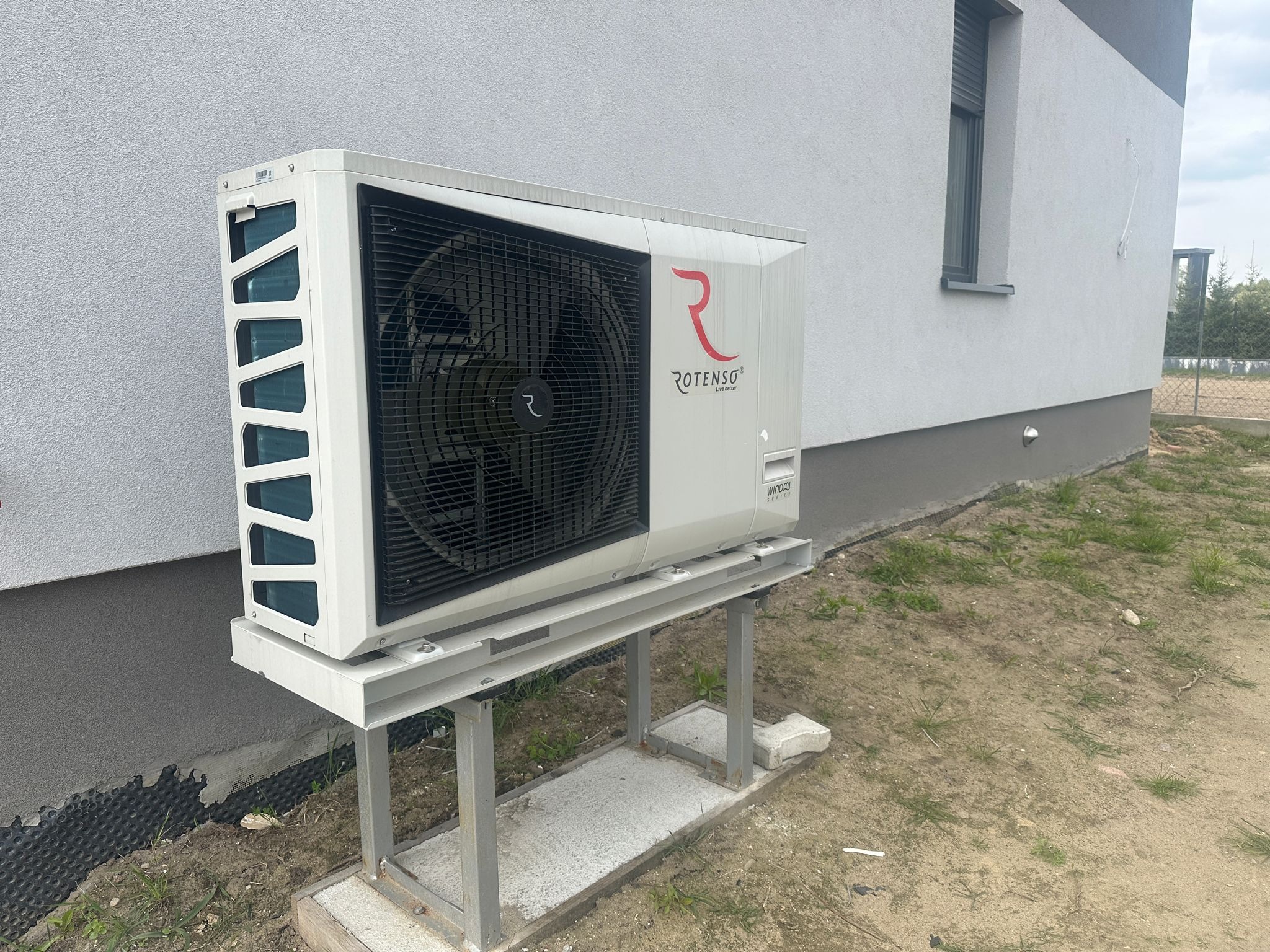 Biała pompa ciepła Rotenso na metalowej konstrukcji przy szarej ścianie budynku. Widoczne logo producenta i fragment okna. Tło: trawa i ogrodzenie.