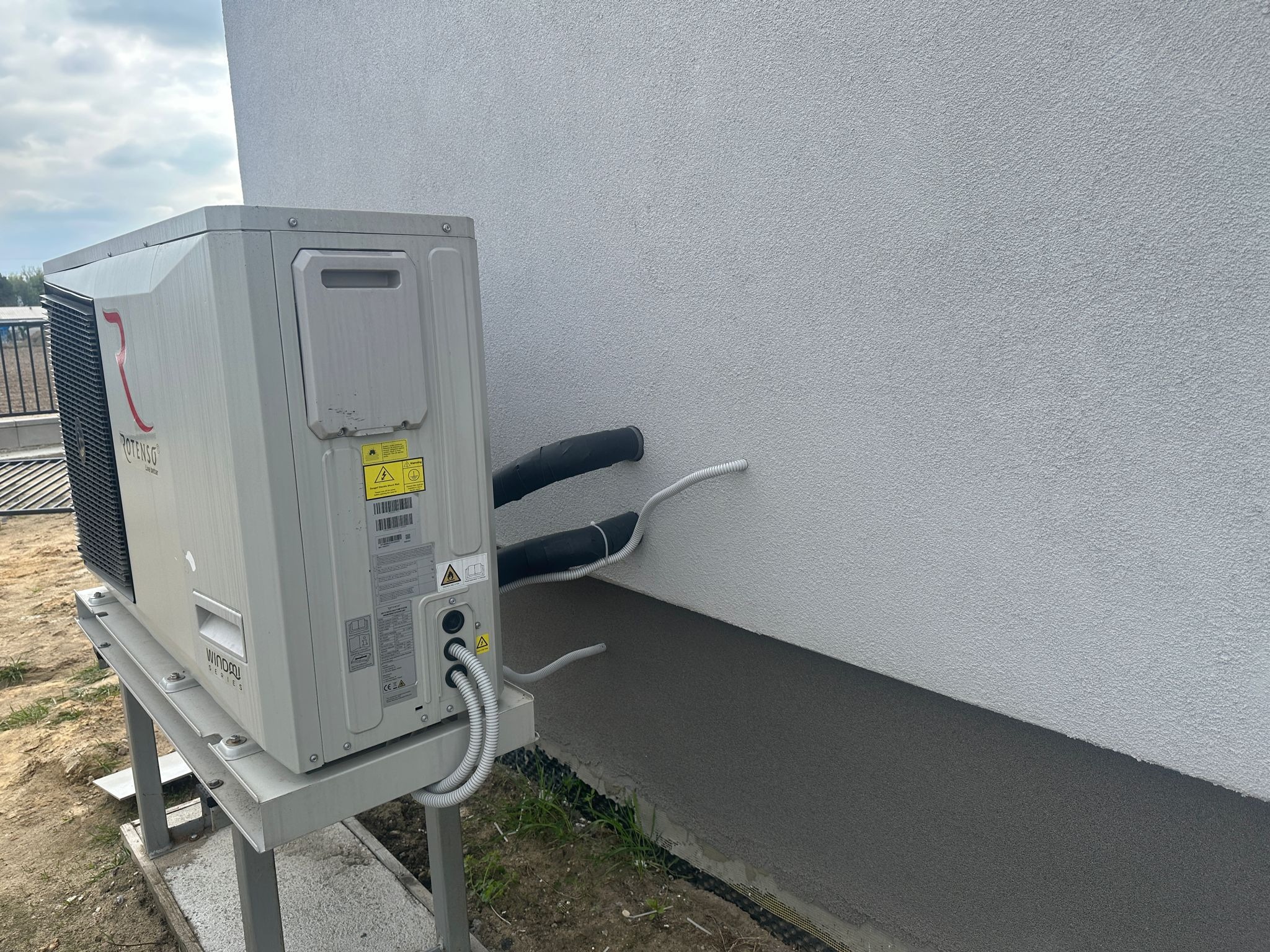 Pompa ciepła Rotenso Windrow Series zamontowana na zewnątrz budynku w Żurominie. Widoczne przyłącza do ściany i metalowa konstrukcja wsporcza.