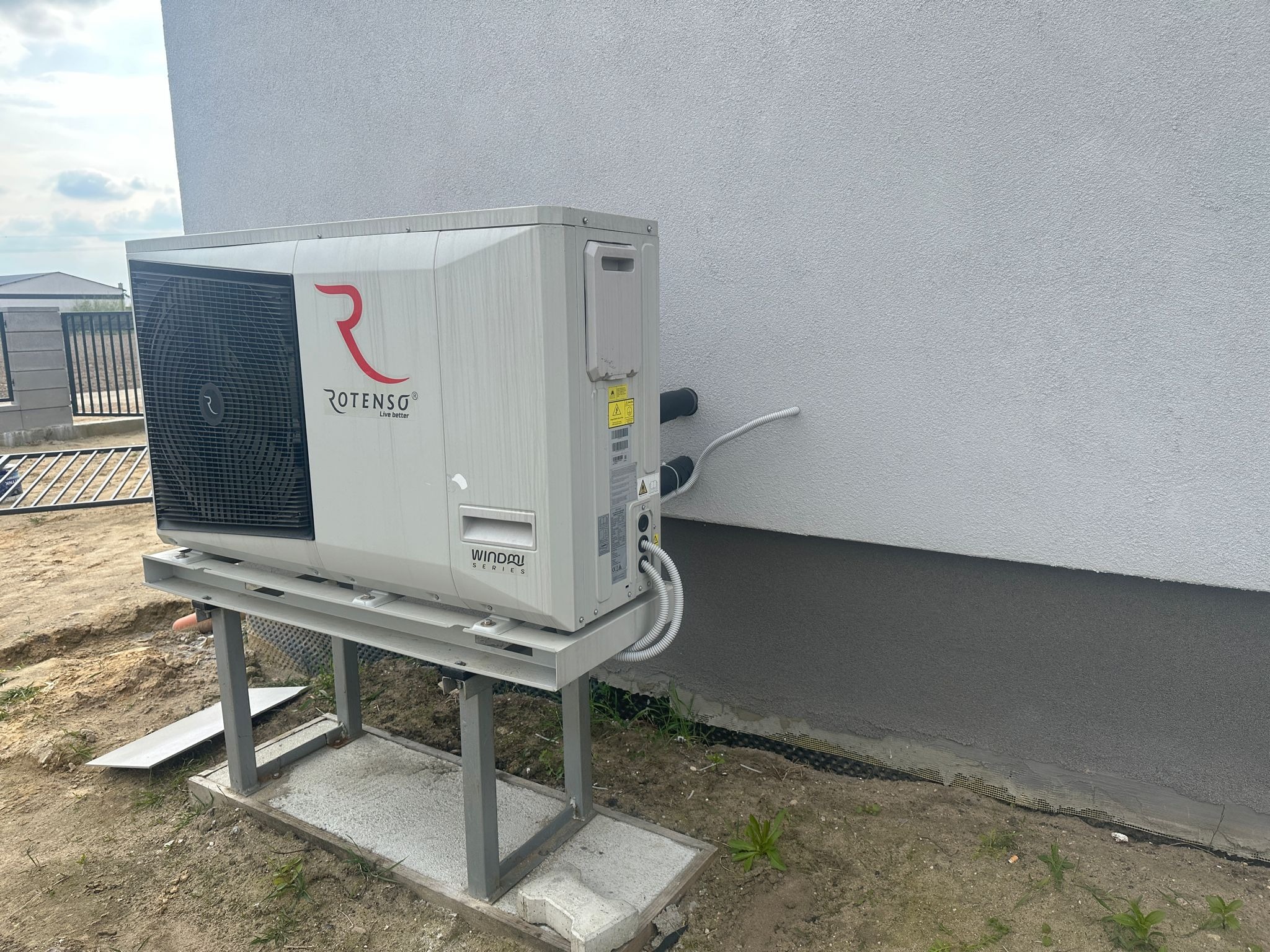 Zewnętrzna jednostka pompy ciepła Rotenso WindAir na metalowej konstrukcji przy jasnej ścianie budynku. Widoczne przyłącza i teren wokół.