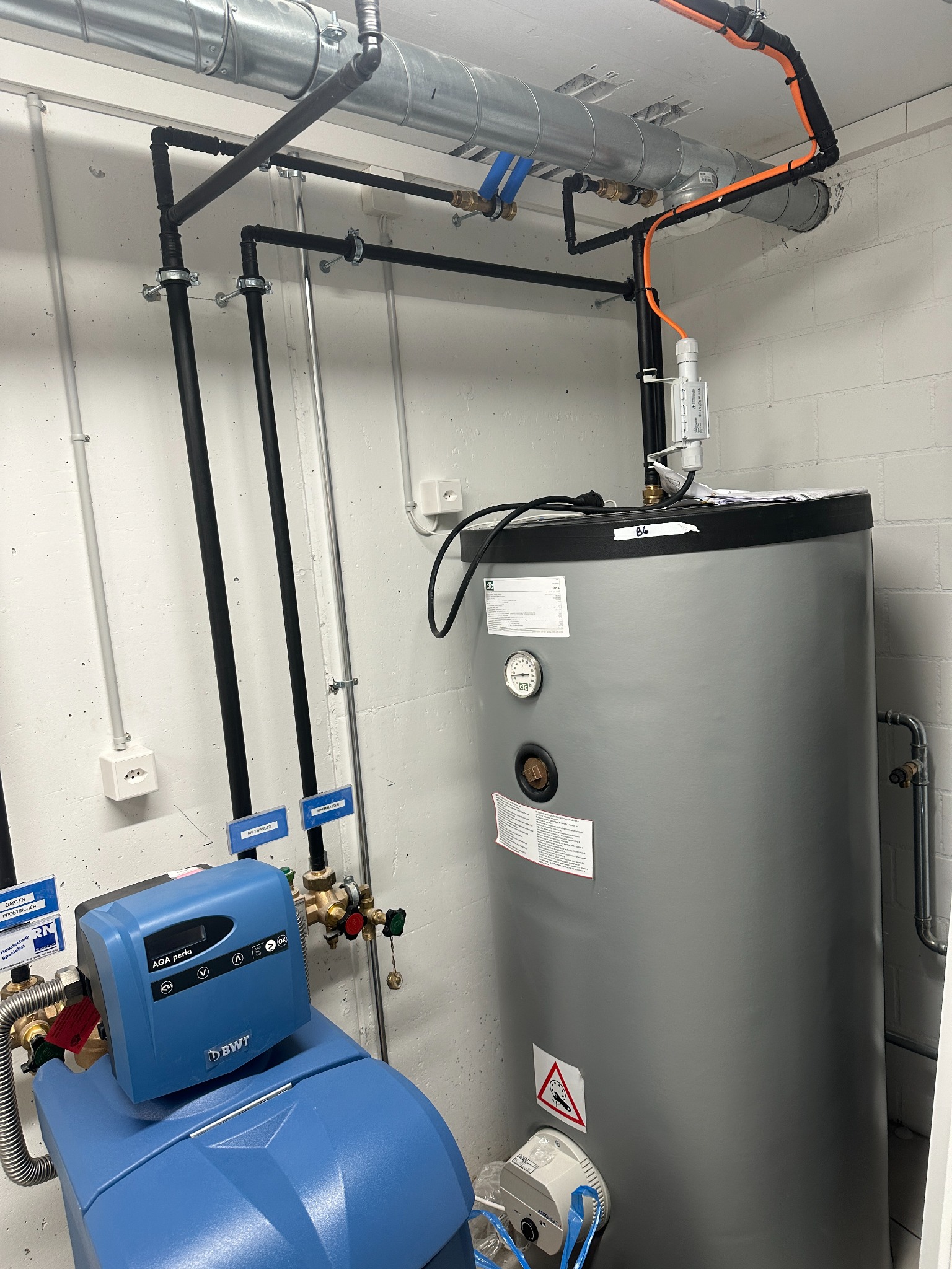 Szary bojler z czarnymi rurami, niebieski zmiękczacz wody BWT AQA Perla i instalacja wentylacyjna w pomieszczeniu technicznym z białymi ścianami.