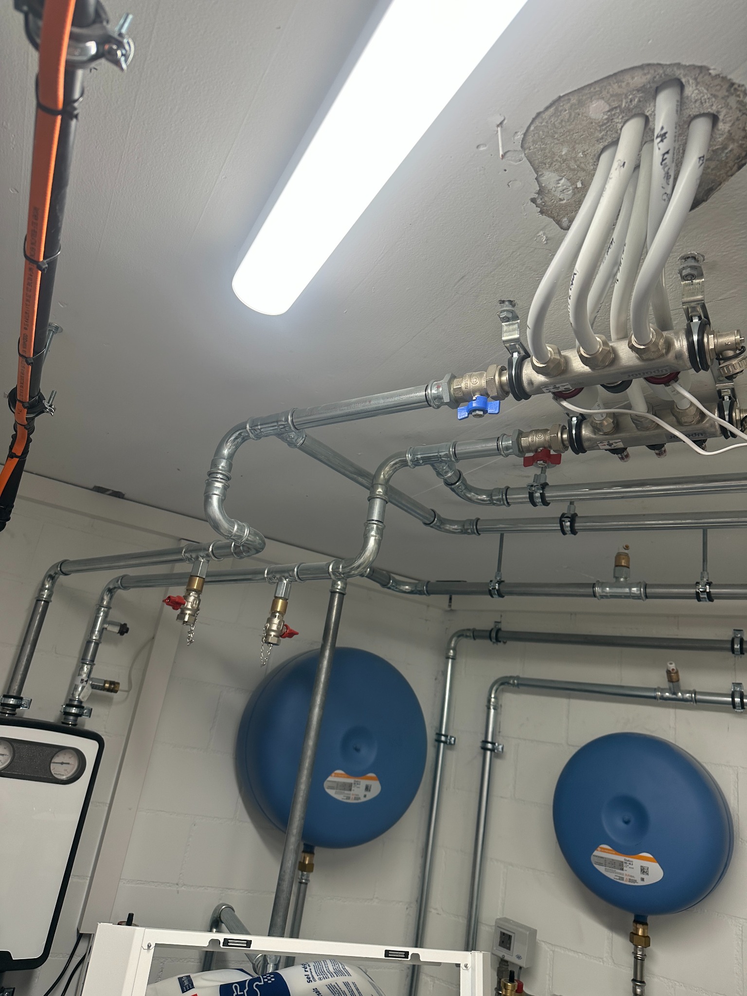 Złożona instalacja grzewcza z rurami, zaworami i zbiornikami wyrównawczymi w pomieszczeniu technicznym. Widoczne elementy kotła i rozdzielacza podłogówki.