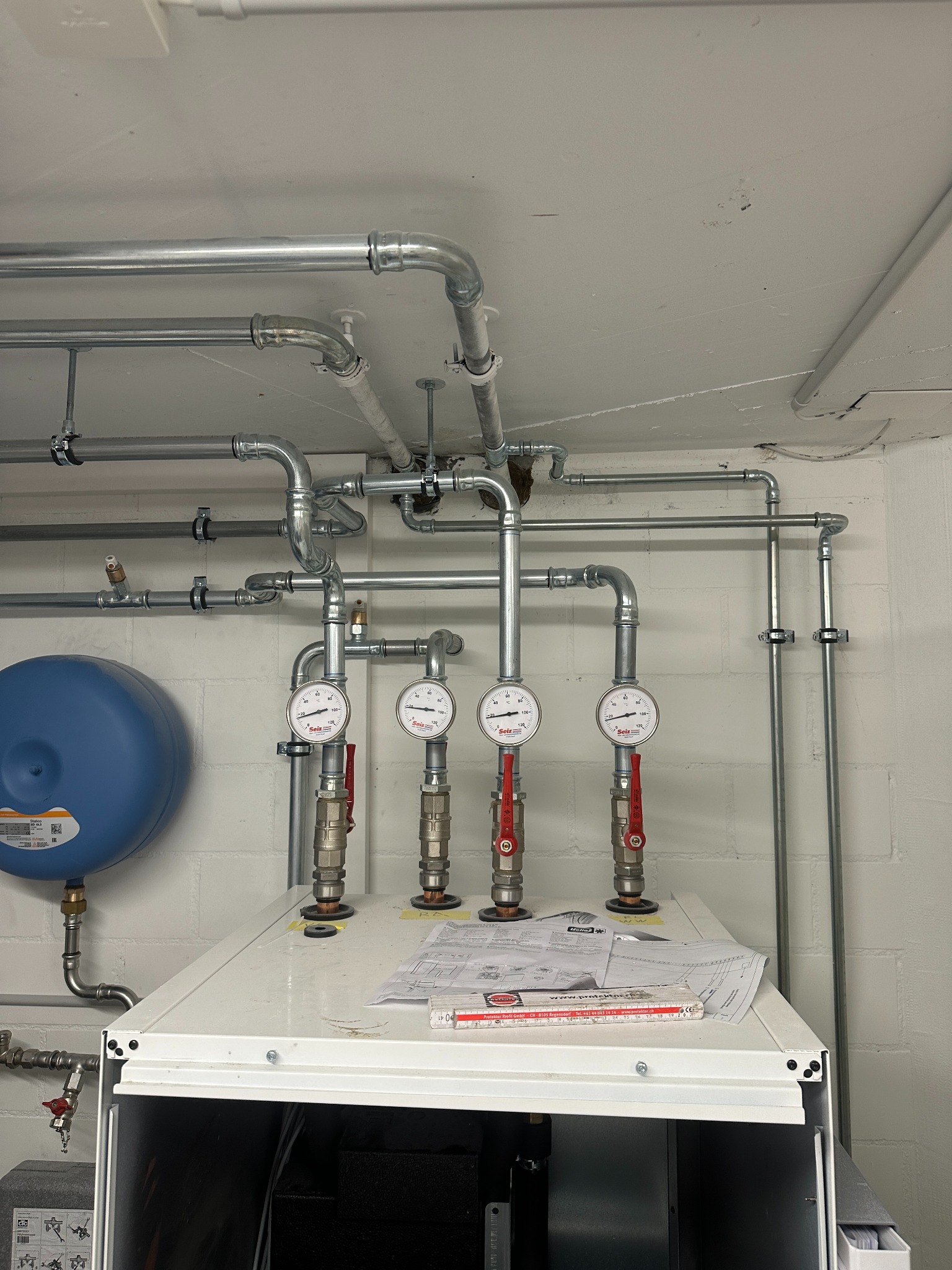 Złożona instalacja grzewcza z rurami, manometrami i zbiornikiem wyrównawczym na tle białej ściany z cegieł. Widoczne złącza i zawory. Na urządzeniu leżą dokumenty i miarka.