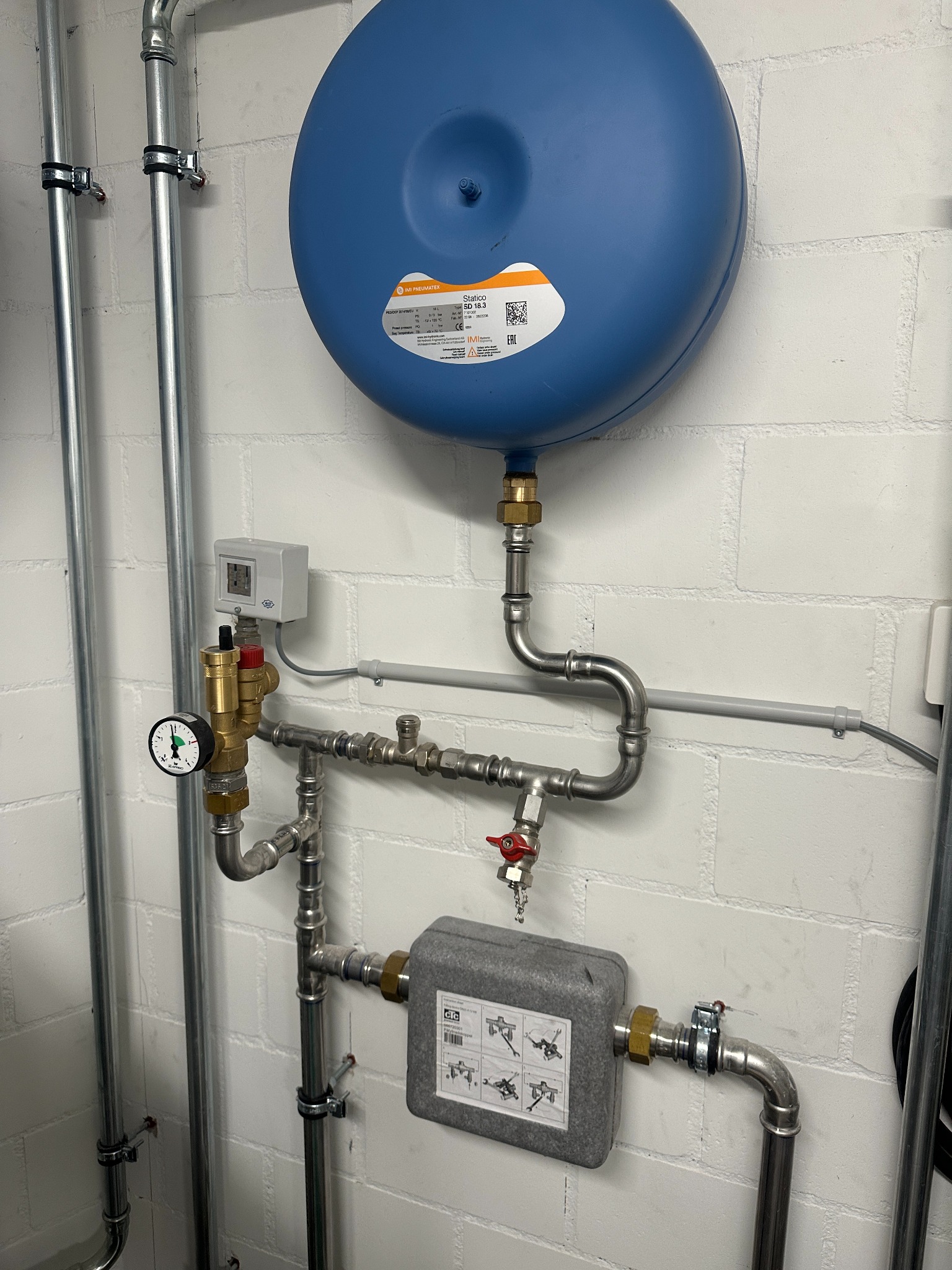 Niebieski zbiornik ciśnieniowy, manometr i zawory na tle białej ściany z cegły. Elementy instalacji grzewczej z rurami ze stali nierdzewnej, widoczne złącza i etykiety.