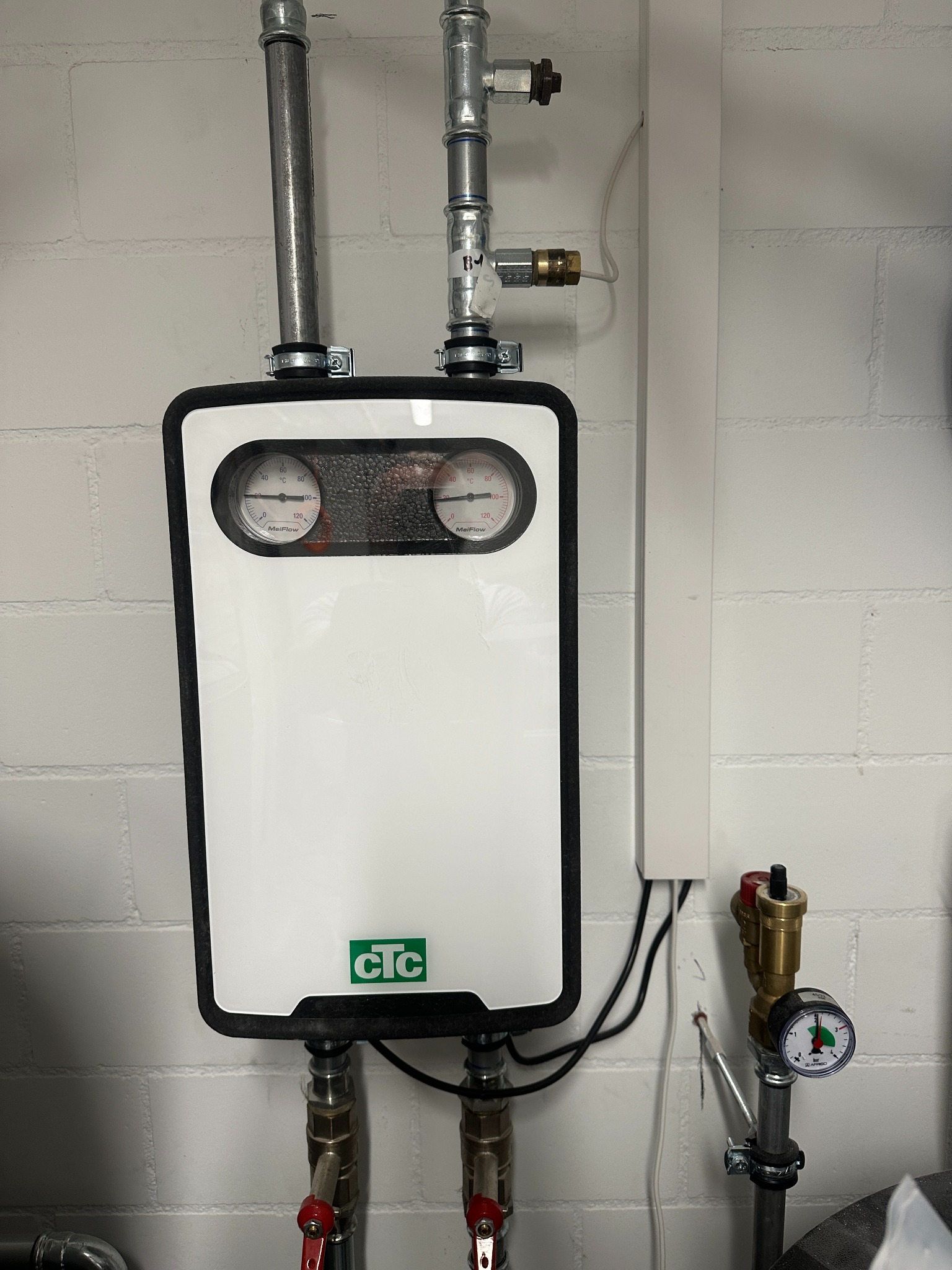 Biała, kompaktowa instalacja grzewcza CTC na ścianie z cegły, z manometrami MelFlow i rurami z zaworami, ukazująca schludny montaż.