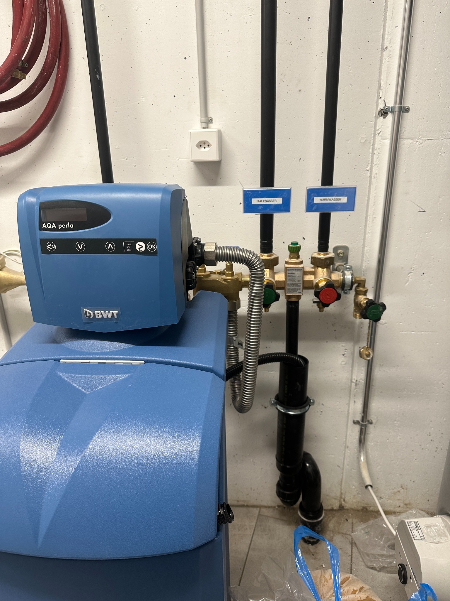 Niebieski zmiękczacz wody BWT AQA Perla z podłączonymi rurami, zaworami i oznaczeniami 'Kaltwasser' i 'Warmwasser' w kotłowni lub pomieszczeniu technicznym.
