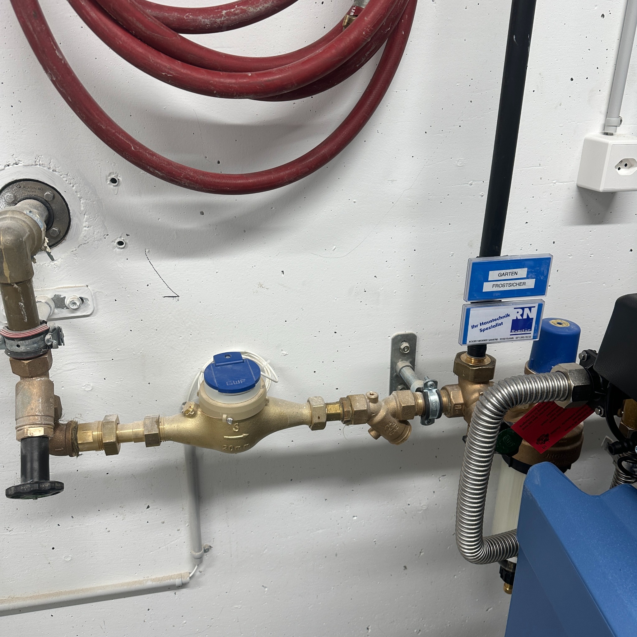 Zamontowany wodomierz z rurami, zaworami i przyłączami na białej, teksturowanej ścianie. Widoczny przewód i etykiety 'Garten' oraz logo firmy instalacyjnej.