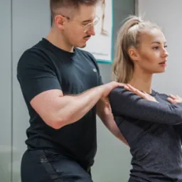 Mężczyzna w okularach, w czarnej koszulce z logo Lacoste, klęczy i masuje górną część pleców blondynki siedzącej na leżance w szarej koszulce na tle plakatu z modelem szkieletu.