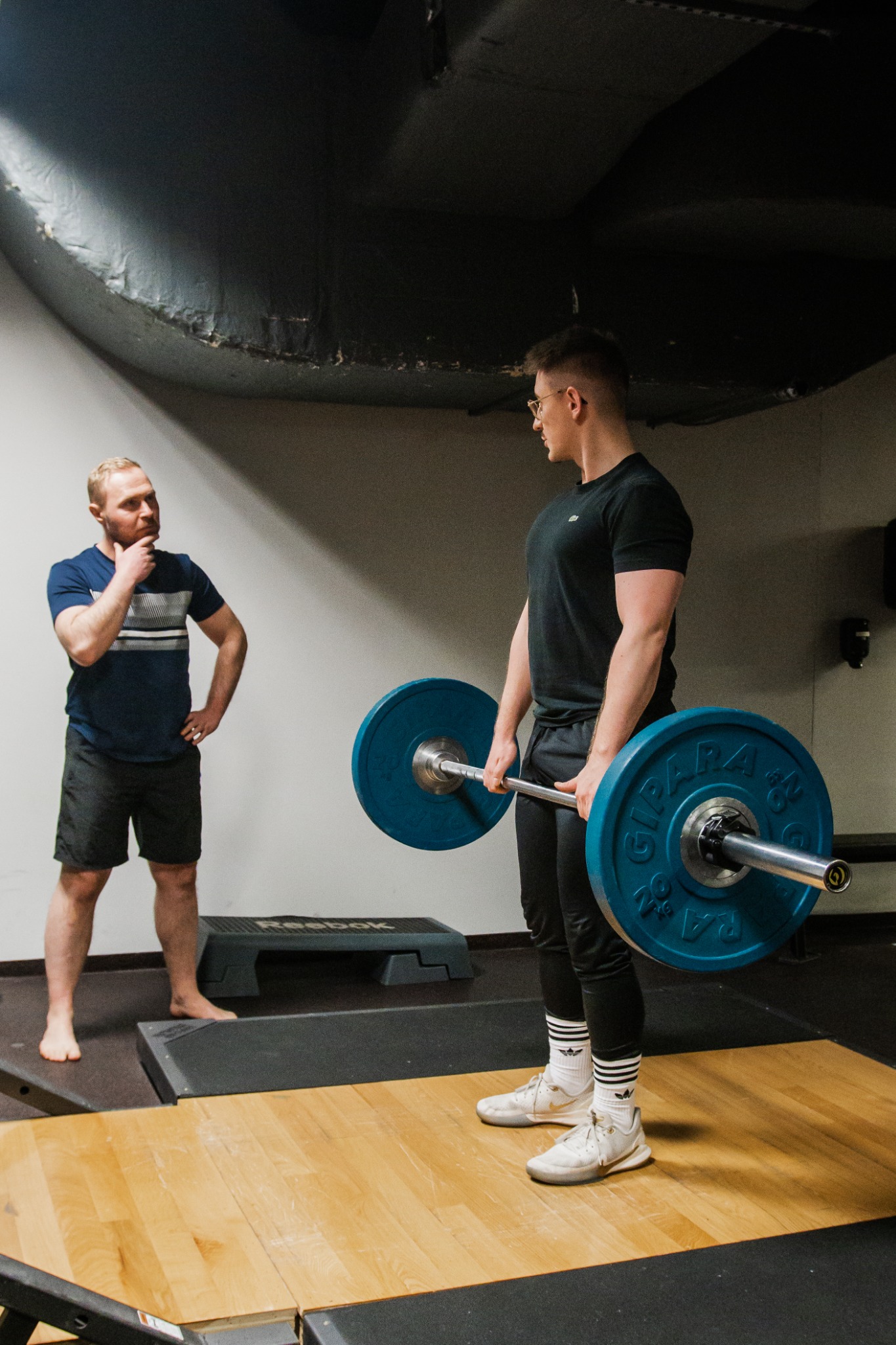 Młody mężczyzna w okularach podnosi sztangę z niebieskimi obciążeniami na drewnianym podeście, podczas gdy drugi mężczyzna obserwuje z założonymi rękami, stojąc na podwyższeniu.
