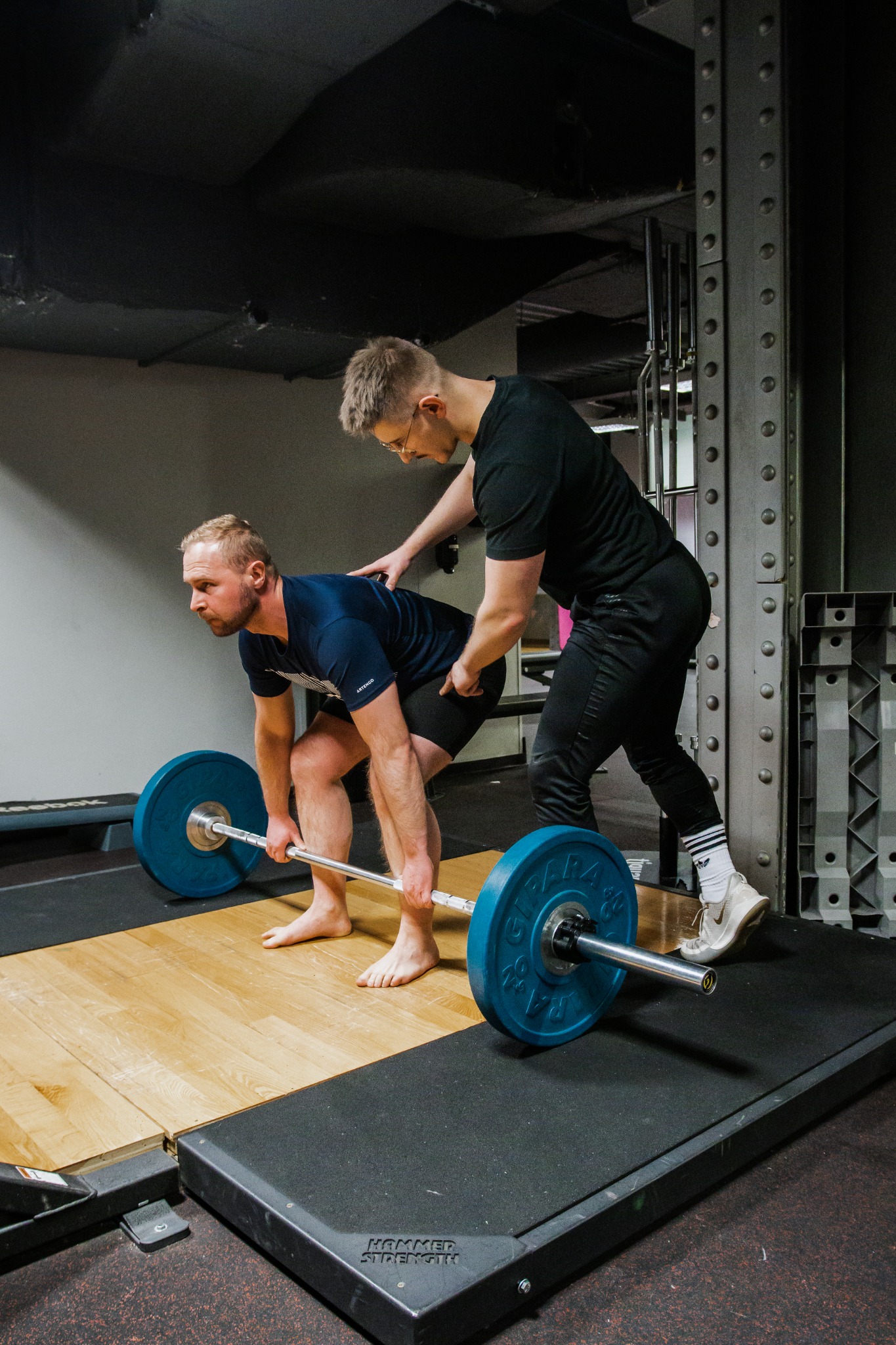 Mężczyzna w niebieskiej koszulce podnosi sztangę z niebieskimi obciążeniami na drewnianej platformie, drugi mężczyzna w czarnym stroju koryguje jego postawę w siłowni.