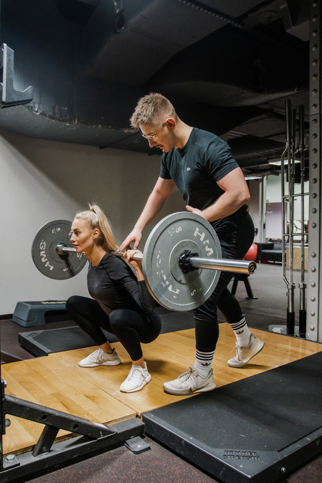 Kobieta wykonuje przysiad ze sztangą pod nadzorem mężczyzny w okularach w siłowni, na platformie z drewnianą podłogą, widoczne obciążenia i logo Hammer Strength.
