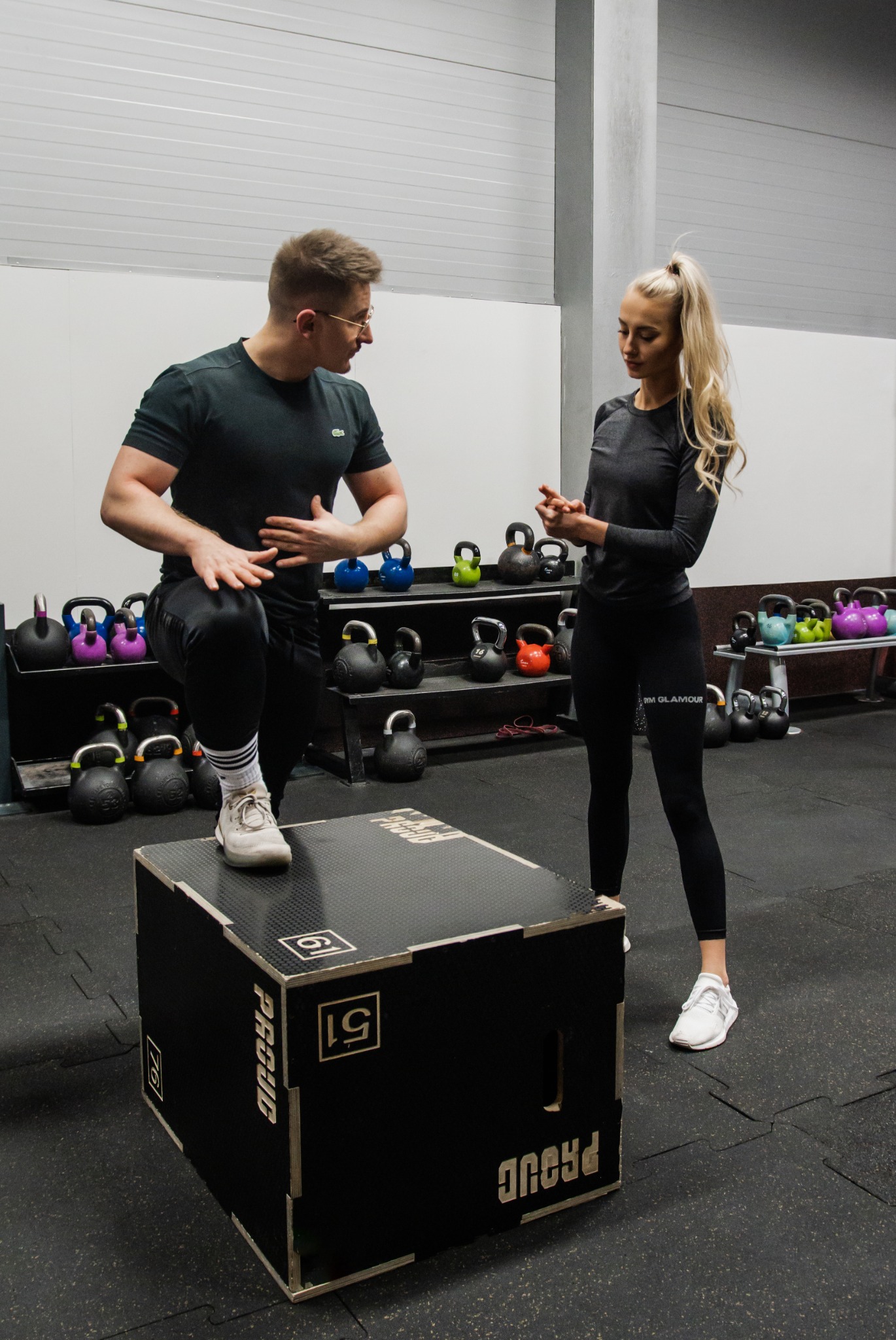 Mężczyzna z okularami demonstruje ćwiczenie na skrzyni, kobieta w sportowym stroju obserwuje, w tle półki z kettlebells