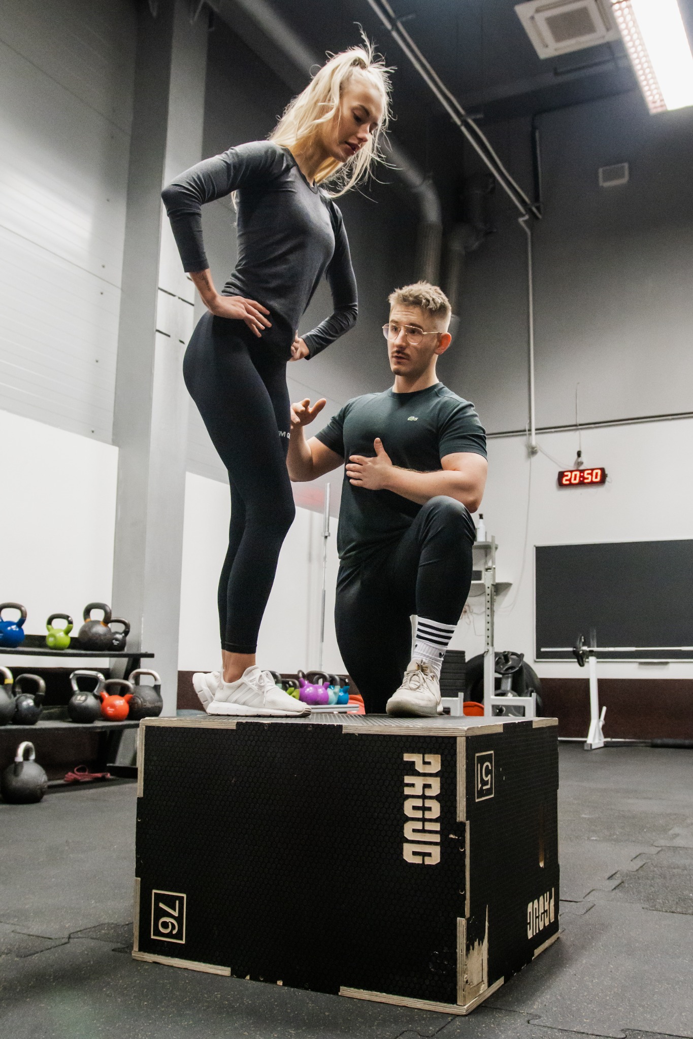 Kobieta w sportowym stroju stoi na skrzyni, mężczyzna w okularach asekuruje, siłownia z kettlebell w tle.