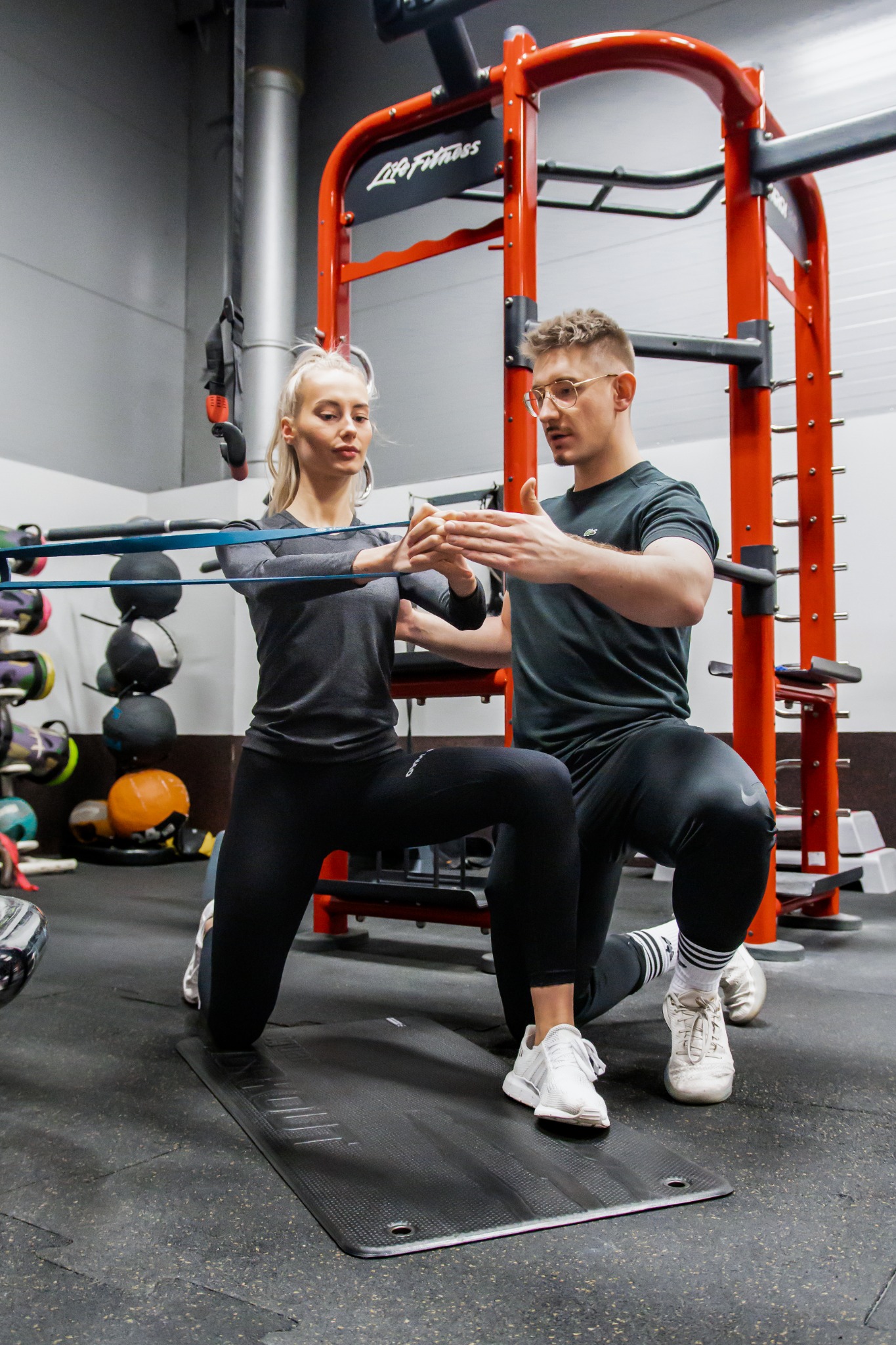 Kobieta klęczy na macie, ćwicząc z gumą oporową pod okiem mężczyzny w okularach, w tle sprzęt do ćwiczeń siłowych marki Life Fitness.