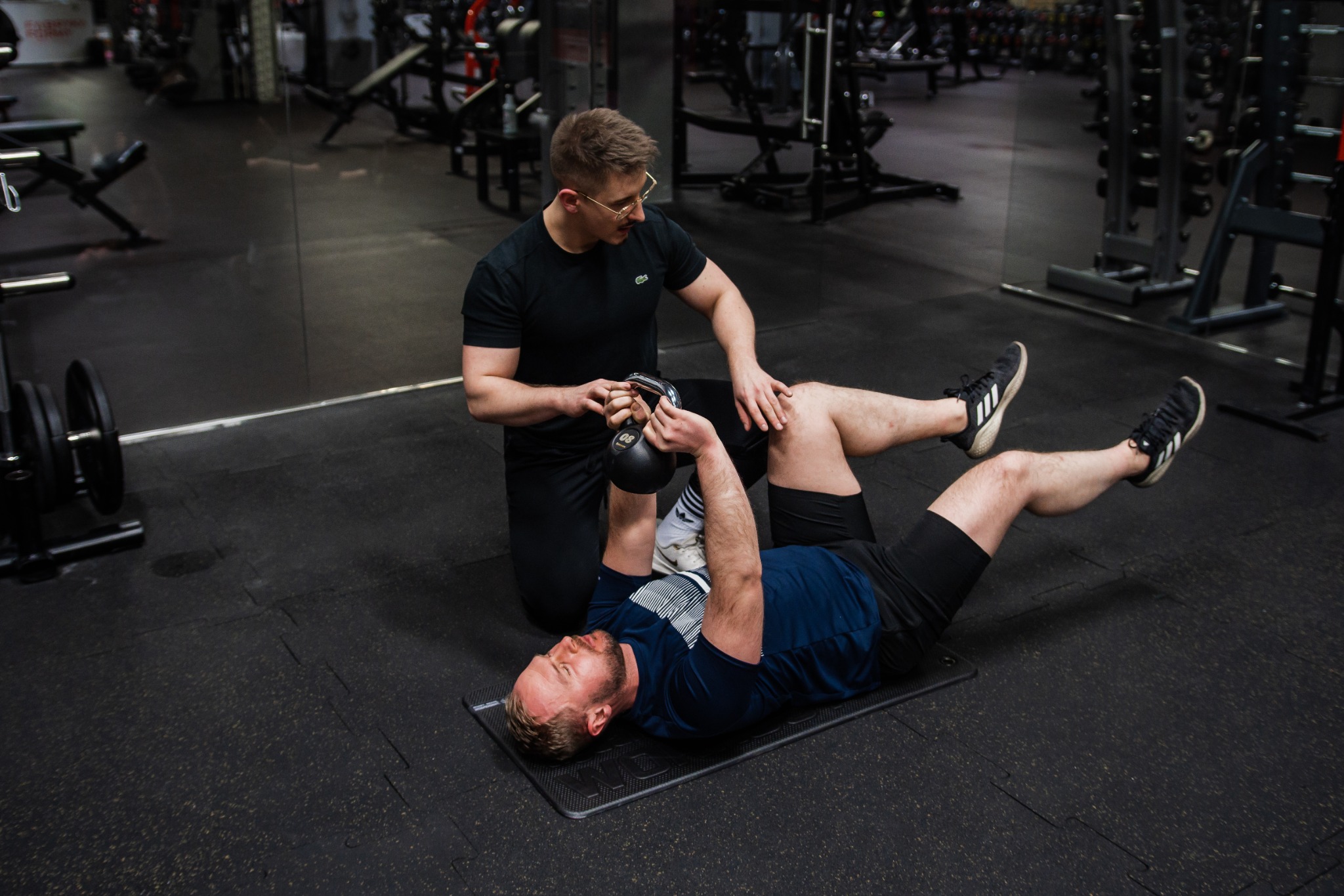 Mężczyzna leżący na macie wykonuje ćwiczenie z kettlebell pod nadzorem trenera w okularach w siłowni.
