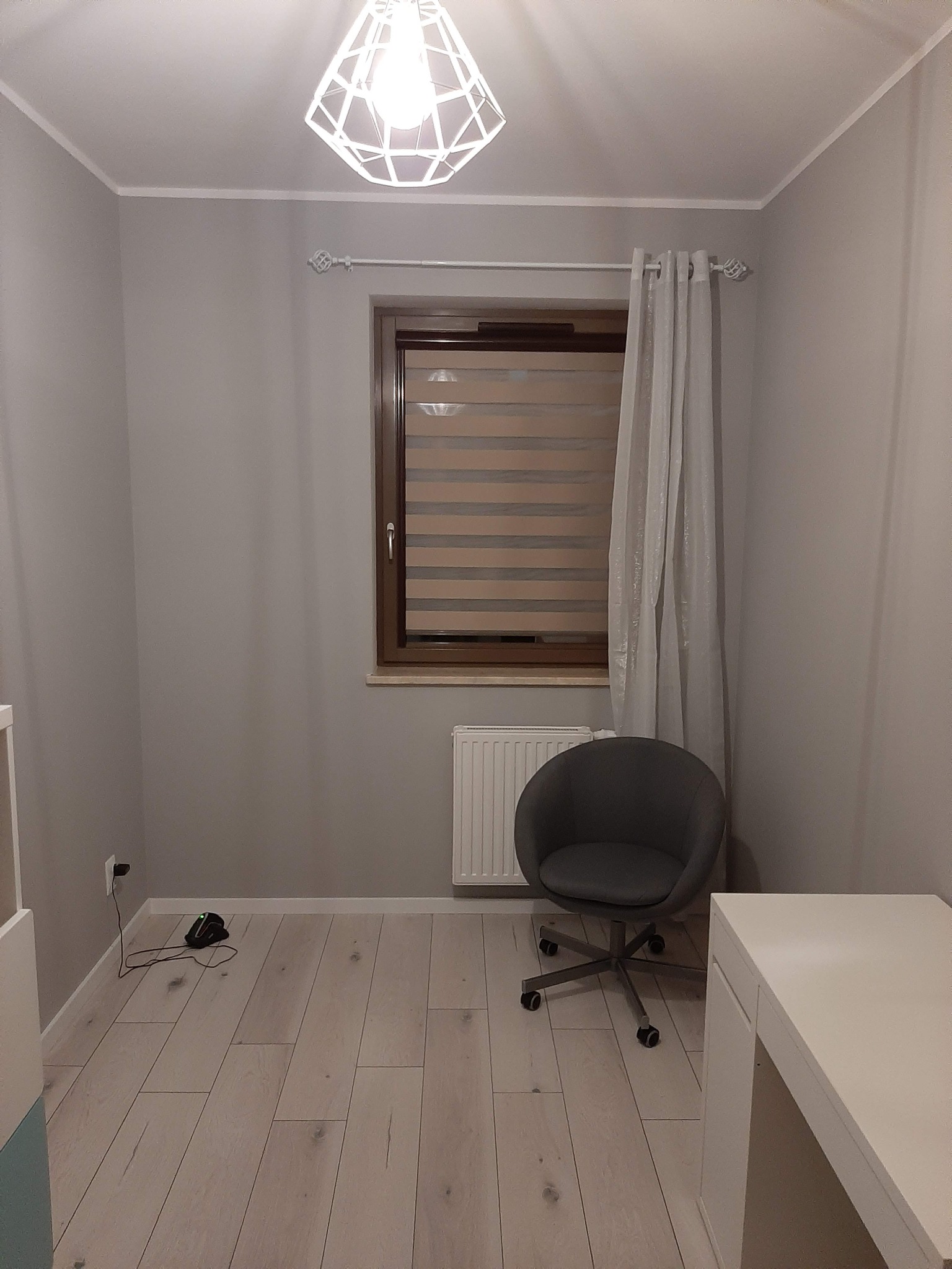 Pokój w szarościach z drewnianą podłogą, okno z roletami dzień-noc, szary fotel na kółkach i białe biurko, oświetlony nowoczesną lampą sufitową.