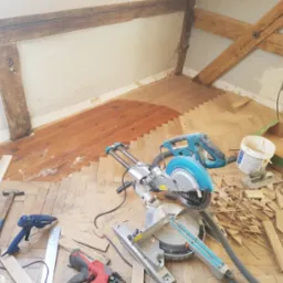 Fragment układania parkietu w jodełkę w pomieszczeniu z drewnianymi belkami stropowymi, widoczne narzędzia: ukośnica, gwoździarka, młotek i pistolet do kleju.