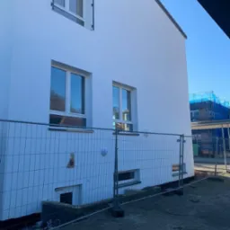 Biała, odnowiona elewacja budynku z trzema oknami, zabezpieczona metalowym ogrodzeniem tymczasowym, z widocznym fragmentem niebieskich rusztowań w tle.