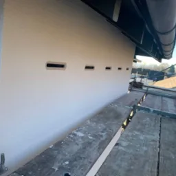 Świeżo otynkowana elewacja domu z małymi otworami wentylacyjnymi, widoczna z rusztowania, w tle zabudowa miejska i błękitne niebo.