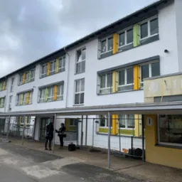 Dwupiętrowy budynek szkoły z białymi ścianami i kolorowymi elementami okiennymi, widoczne rusztowania i dwie osoby przed wejściem.