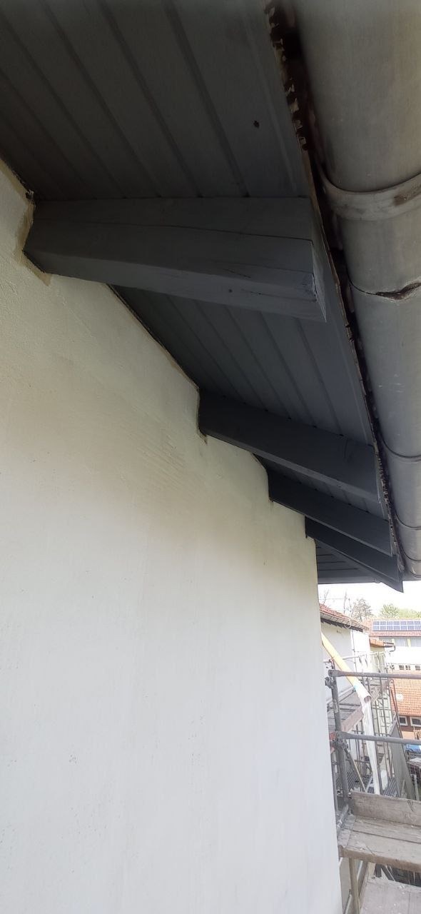 Świeżo otynkowana elewacja budynku z widocznymi elementami drewnianymi pod dachem pomalowanymi na szaro, fragment rynny i rusztowania w tle.