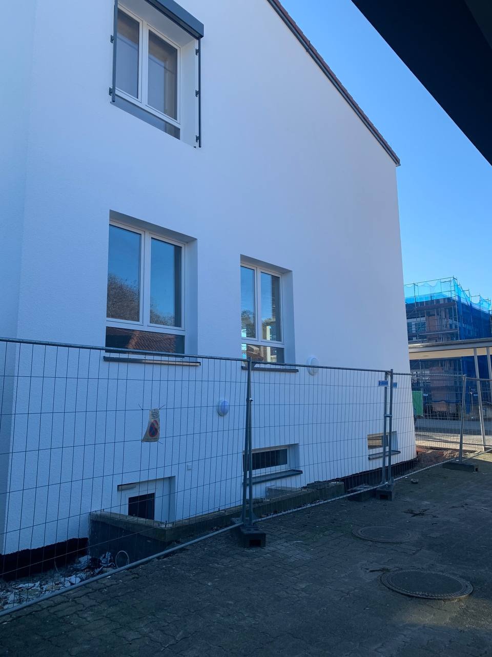 Biała, odnowiona elewacja budynku z trzema oknami, zabezpieczona metalowym ogrodzeniem tymczasowym, z widocznym fragmentem niebieskich rusztowań w tle.