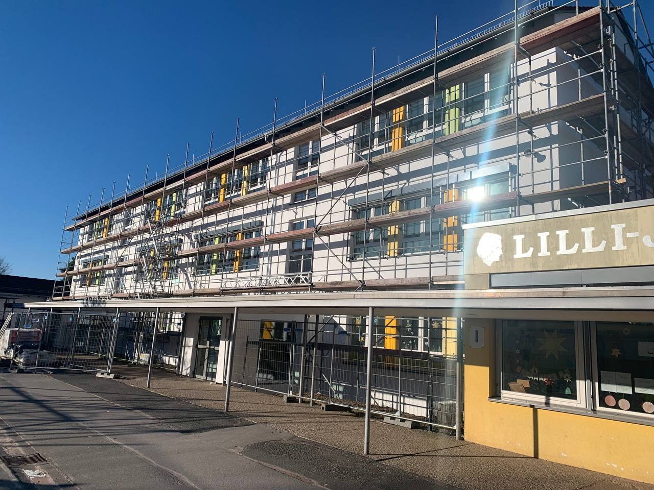 Trzypiętrowy budynek szkolny z rusztowaniami, przygotowywany do prac elewacyjnych, widoczne kolorowe elementy okien i tablica z nazwą 'Lilli-'.