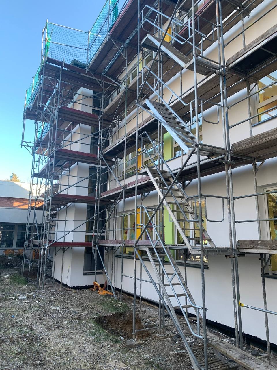 Budynek w trakcie ocieplania elewacji, widoczne rusztowania, częściowo otynkowana ściana, okna w kolorze żółtym i zielonym, wykop w ziemi przy budynku.